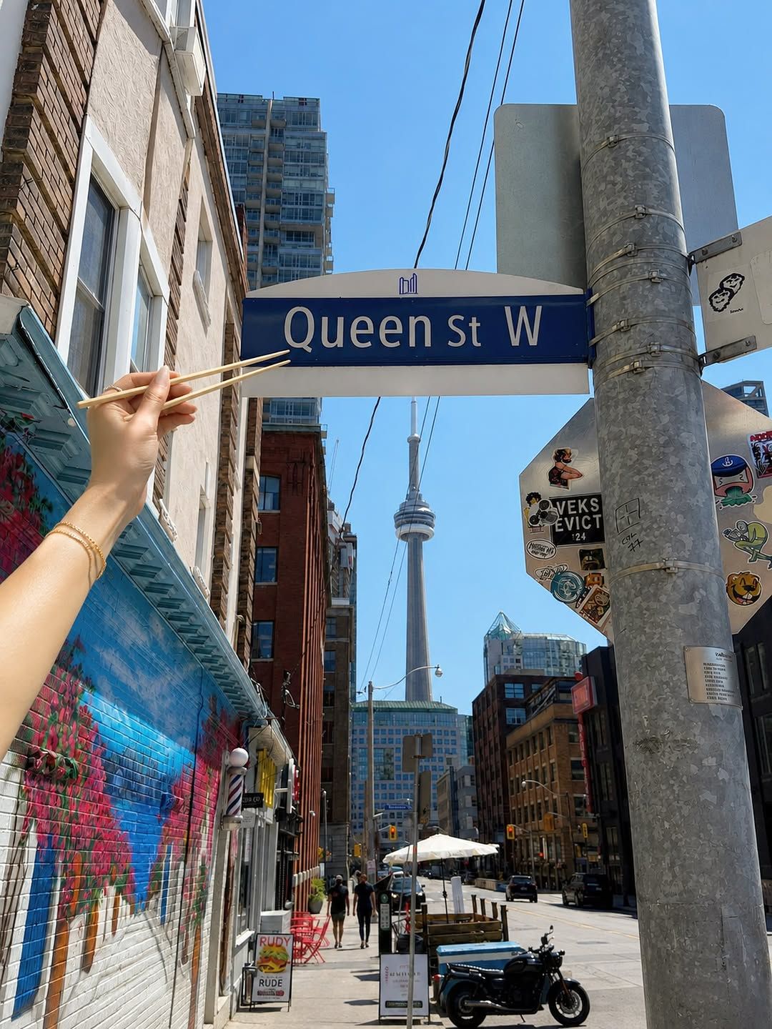 Chopsticks pointing at a Queen St W street sign with the CN Tower behind, posted by @kajikencanada — location teaser, not a Kajiken storefront